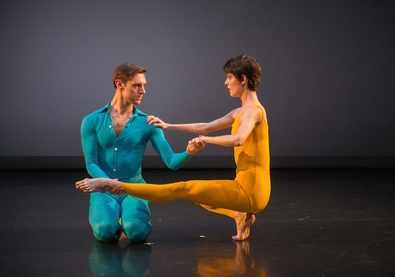 Benny Olk, in a bright blue unitard with buttons up the front and collar, kneels and holds Vanessa Knouse's hand and ankle as she assumes a squated position in which her left leg extends in front of her.